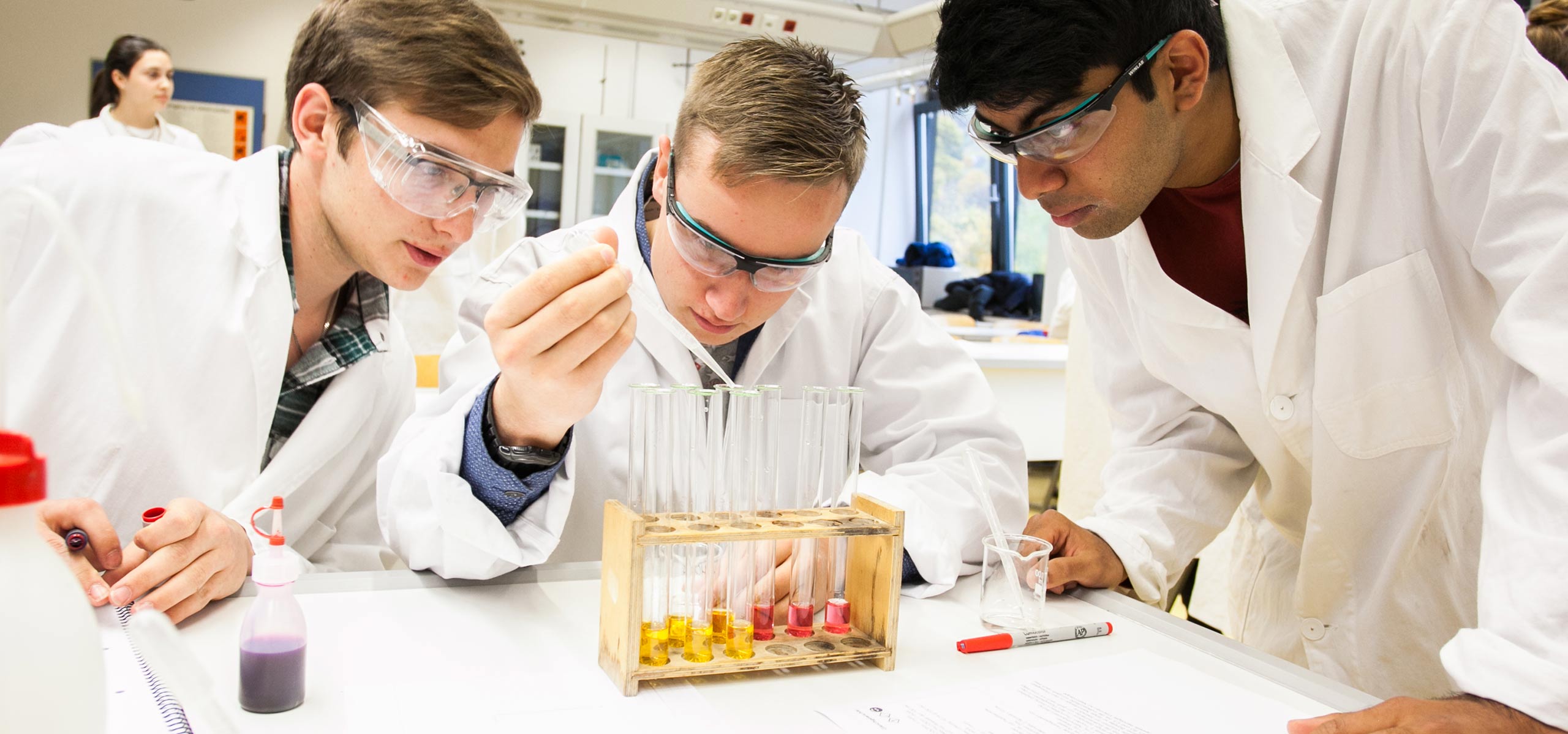 Ein Lehrer zeigt drei Schülern im Chemiesaal. Auf dem Tisch stehen weitere Utensilien aus dem Chemieunterricht. Alle Personen tragen Schutzkleidung und beobachten das Experiment.